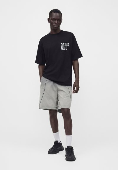 Άνδρας που φοράει μαύρο oversized t-shirt με γραφικό κείμενο, γκρι σορτς μέχρι το γόνατο με ραφές, λευκές κάλτσες και μαύρα αθλητικά παπούτσια, στέκεται μπροστά σε λευκό φόντο.