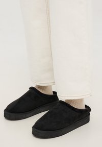 Pantofola nera in morbido suede con interno in pelliccia, dotata di punta arrotondata e suola piatta in gomma, abbinata a pantaloni di colore chiaro.