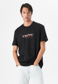 Black cotton t-shirt featuring a red and white graphic logo "tommy JENS" on the front. Short sleeves and a round neckline. Minimalist design.