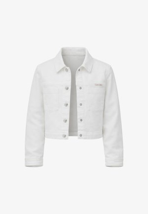 Giovane ragazza con capelli castani, giacca di jeans bianca, camicia bianca, jeans larghi azzurri chiari e sandali neri, in piedi con una mano in tasca.