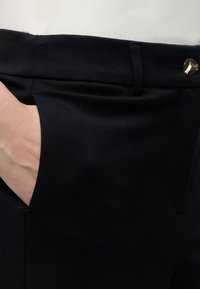 Main dans la poche d’un pantalon habillé noir avec passants de ceinture et bouton doré, associé à un haut blanc.