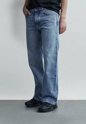 Personne portant un jean bleu à coupe décontractée avec le logo « Off-White » près de la poche, des baskets noires et une chemise noire, debout devant un fond uni.