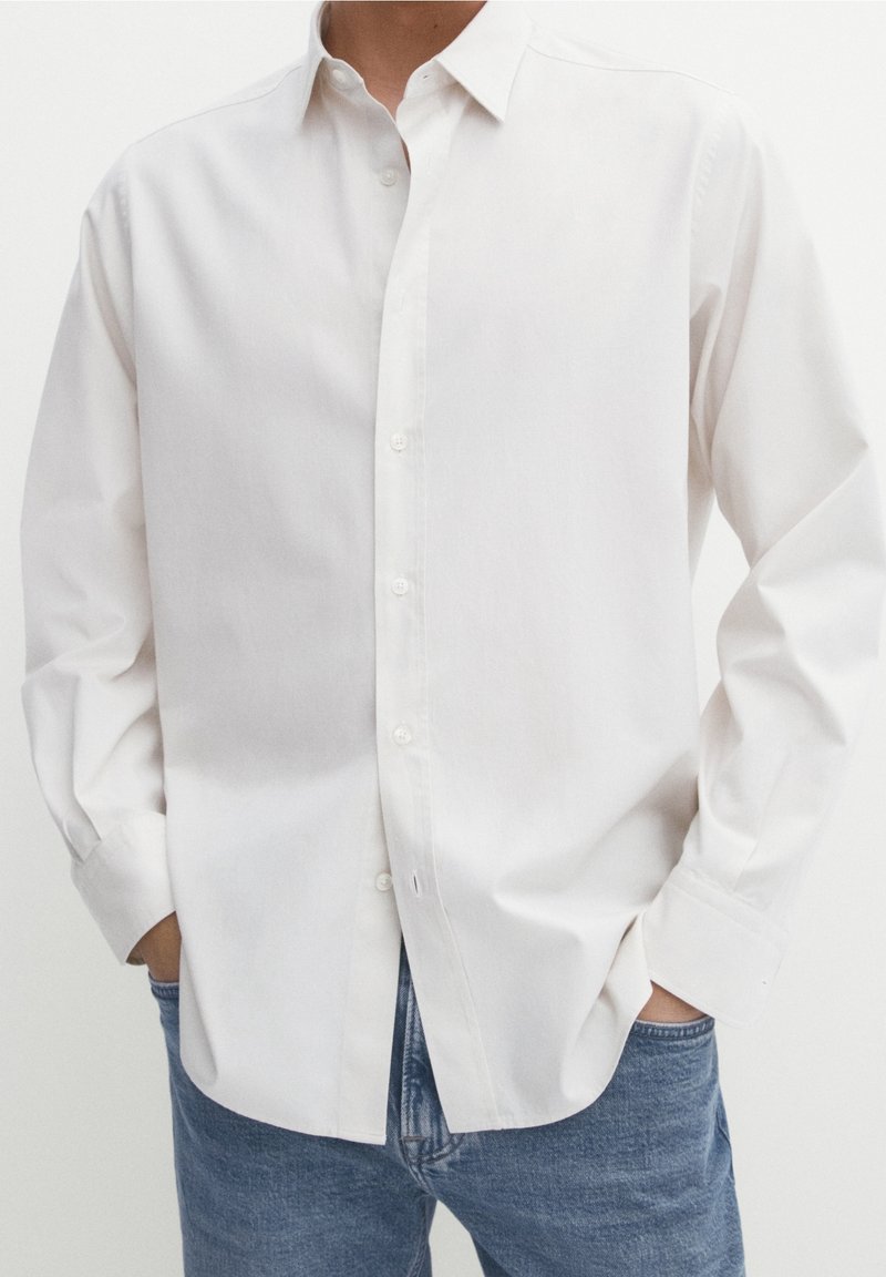 Personne portant une chemise blanche à manches longues avec col, non rentrée sur un jean en denim bleu, mains dans les poches.