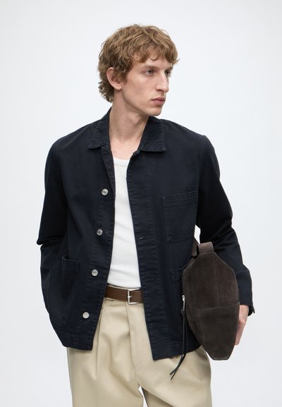 Jeune homme aux cheveux blonds bouclés portant une veste noire boutonnée, une chemise blanche, un pantalon beige, tenant un sac en daim brun foncé.