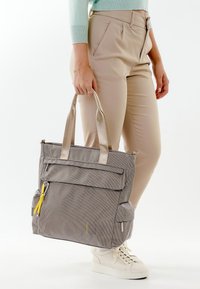 Persona che indossa pantaloni beige e scarpe da ginnastica bianche, che tiene per le maniglie una grande borsa grigia con cerniere gialle.