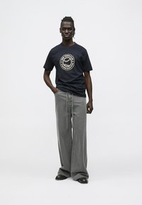 Tamsiai mėlynas marškinėliai su apvaliu teniso klubo logotipu; laisvo kirpimo pilkos kelnės; dėvimos su juodais batais; lygus audinys ir laisva siluetas.