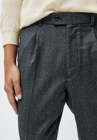 Graue Wollhose mit feinem Muster, ausgestattet mit Knopfverschluss, Falten und glatter Textur, kombiniert mit einem hellcremefarbenen Pullover.