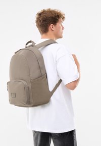 Rucsac din țesătură gri, cu o formă rotunjită, buzunar frontal cu fermoar și bretele umplute. Buzunar lateral din plasă și accente texturate.