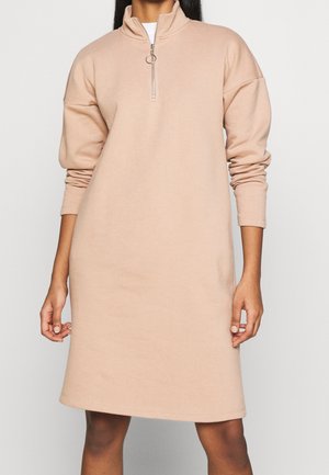 Femme portant une robe beige à manches longues, longueur genou, col montant et fermeture éclair avec anneau argenté, debout devant un fond uni.
