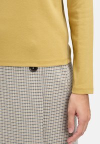 Żółta bluzka z długim rękawem o gładkiej fakturze, noszona na wierzchu w kratkę spódnicy z ciemnobrązowym detalem w postaci guzika.