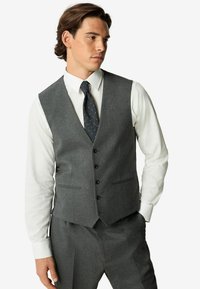Gilet de costume gris en tissu mélangé de laine, avec un décolleté en V, six boutons et deux poches avant. Porté par-dessus une chemise blanche.