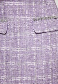 Lila Tweedstoff mit einem strukturierten Muster aus weißen und goldenen Fäden. Zwei Vordertaschen mit dekorativen silbernen Akzenten.