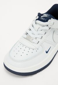 Sneaker en cuir blanc avec des accents navy, présentant des perforations sur le bout, des lacets plats et un détail de logo argenté sur la languette.