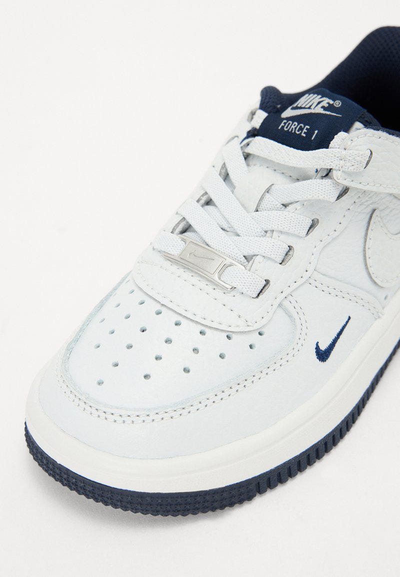 Weiße Leder-Sneaker mit marineblauen Akzenten, ausgestattet mit Perforationen an der Zehenpartie, flachen Schnürsenkeln und einem silbernen Logodetail auf der Zunge.