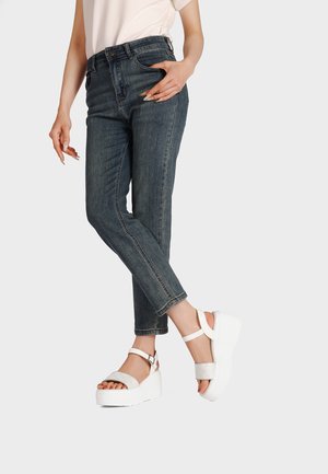 Donna che indossa jeans slim fit scuri e sandali con zeppa bianchi, in piedi con una gamba leggermente sollevata su uno sfondo uniforme.