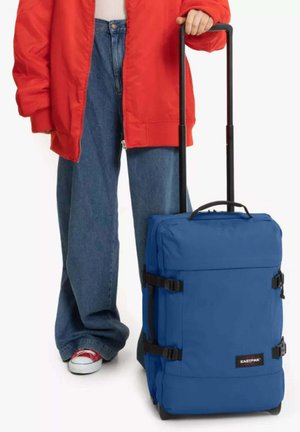 Personne en veste rouge et jean tenant la poignée d'une valise à roulettes Eastpak bleue, debout sur fond blanc.