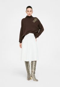 Brązowy sweter z dzianiny z wysokim kołnierzem, biały spódnica A-line oraz srebrne botki w jadowity wzór węża do kolan z blokowym obcasem. Udekorowany ozdobami.