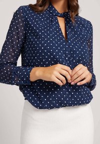 Blouse bleu marine avec manches longues transparentes et encolure goutte d'eau, présentant un motif à pois blancs et bleus, assortie à une jupe blanche côtelée.