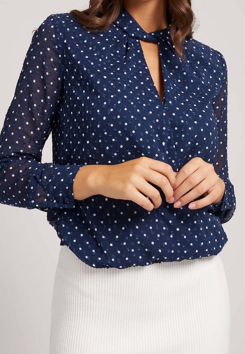Blouse bleu marine avec manches longues transparentes et encolure goutte d'eau, présentant un motif à pois blancs et bleus, assortie à une jupe blanche côtelée.