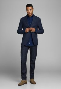 Blazer blu navy sopra una camicia a pois, abbinato a jeans di denim scuro e stivali in suede beige. Vestibilità su misura con spalle strutturate e bottoni.