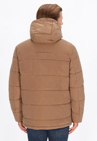Jachetă puf din brown cu o glugă mare, design matlasat și o croială relaxată. Textură netedă cu cusături orizontale, asortată cu blugi din denim albastru.