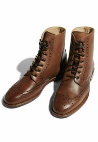 Stivali alla caviglia in pelle marrone con dettagli a brogue, dotati di design con lacci, punta rotonda e lacci neri a contrasto. Finitura liscia e lucida.