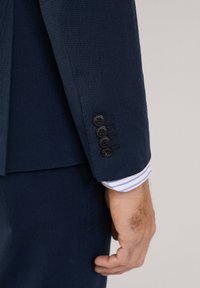 Navyfarbener, strukturierter Blazer mit Ärmel und vier schwarzen Knöpfen, der darunter einen kontrastierenden gestreiften Umschlag in Weiß und Blau zeigt.