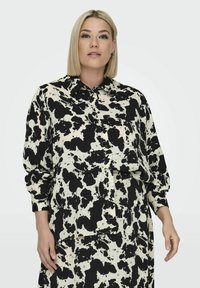 Sort og hvid prikket skjorte lavet af et letvægtsstof. Den har en krave, knaplukning foran og lange ærmer med løst fit.