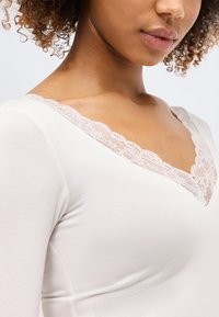 Junge Frau mit lockigem Haar, die ein weißes Oberteil mit Spitzenbesatz entlang des V-Ausschnitts trägt, dargestellt von Hals bis Brust.