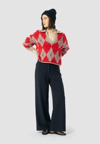Czerwony i beżowy sweter w romby z dekoltem w serek, połączony z czarnymi szerokimi spodniami i czarnymi siateczkowymi balerinami. Modelka nosi czarną czapkę.