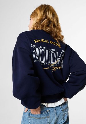Frau mit welligem blondem Haar, die einen dunkelblauen Red Bull Racing Sweatshirt und hellblaue Jeans trägt, die Hände in den Gesäßtaschen, seitlich stehend.