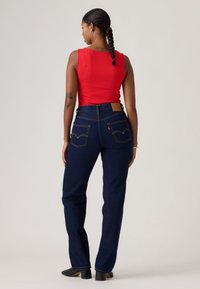 Γυναίκα με πλεγμένα μαλλιά στέκεται πλάτη φορώντας κόκκινο αμάνικο τοπ, σκούρα μπλε τζιν με ίσια γραμμή και μαύρα παπούτσια με τακούνι.