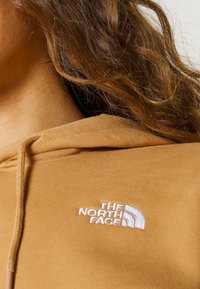 Gros plan d'une personne portant un sweat à capuche beige avec le logo The North Face brodé en blanc sur la poitrine. Cheveux bruns ondulés visibles.