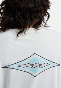 Persona con cabello rizado y oscuro vista desde atrás, que lleva una camisa blanca con un diseño abstracto de diamantes en azul y negro.