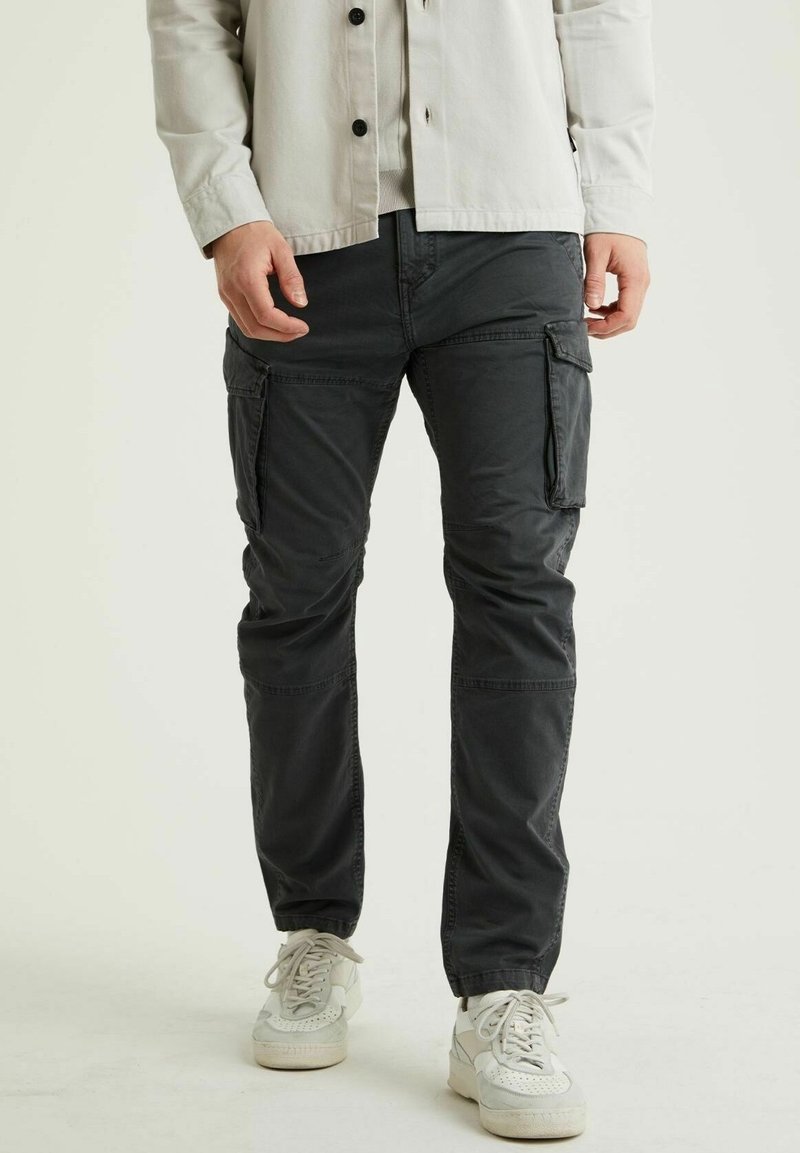 Homme portant un pantalon cargo gris foncé avec des poches latérales et des baskets blanches, debout devant un fond clair uni.