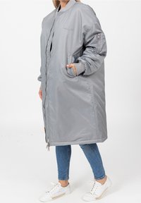 Femme portant une longue veste bomber oversize gris clair avec fermeture éclair à l'avant, poignets côtelés, poches latérales, jean skinny bleu et baskets blanches.