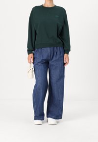 Sweatshirt en coton vert foncé avec col rond, manches longues et logo discret. Associé à un jean large bleu et des baskets blanches.