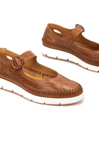 Chaussures en cuir marron avec un design perforé, détail de boucle circulaire et semelle en caoutchouc blanc. Présente un style décontracté avec des textures douces.
