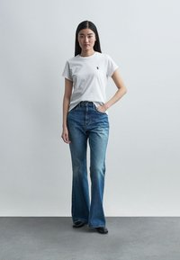 Jonge vrouw met lang donker haar, gekleed in een wit T-shirt en wijde blauwe jeans, staand tegen een effen grijze achtergrond.