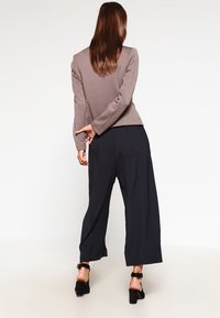 Femme aux longs cheveux bruns vue de dos, portant une veste texturée taupe, un pantalon large bleu marine et des sandales noires à talons blocs avec brides à la cheville.