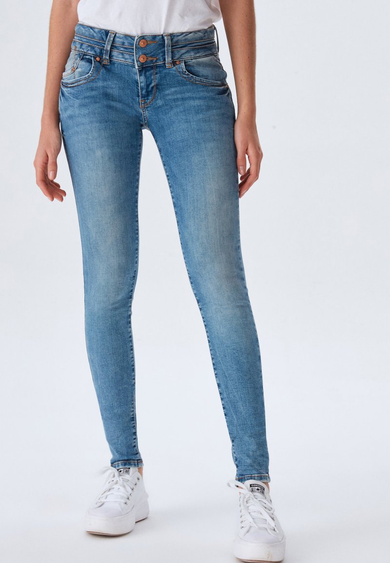 Jeans de mezclilla azul ajustados con un acabado desgastado, cierre de doble botón y detalles cosidos en los bolsillos frontales, combinados con zapatillas blancas.