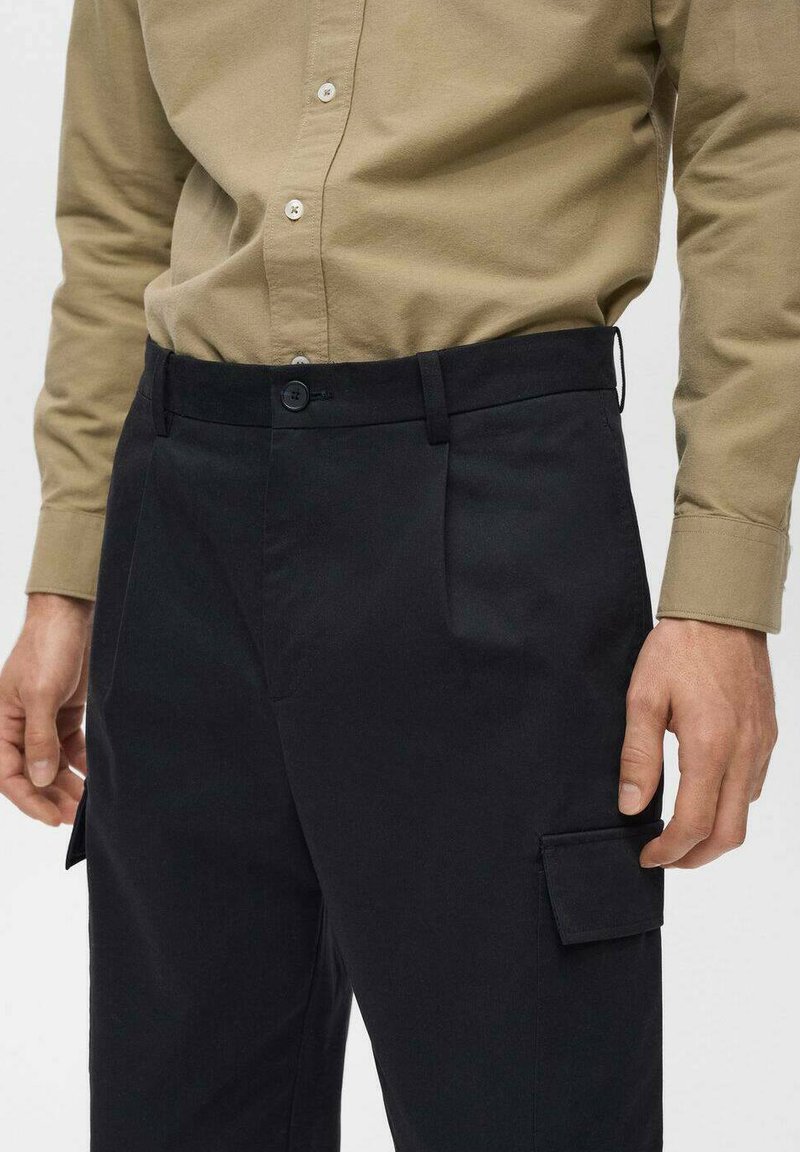 Schwarze, maßgeschneiderte Cargo-Hosen mit glatter Textur, ausgestattet mit Seitentaschen und einem Knopfverschluss am Bund, getragen mit einem khakifarbenen Hemd.