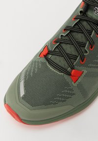 Baskets de sport vertes avec tige en mesh et lacets noirs. Présente des accents rouges, des motifs texturés et une semelle extérieure en caoutchouc durable.