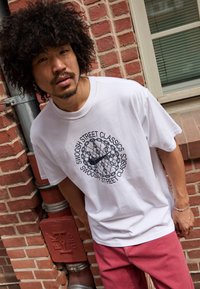 Witte katoenen t-shirt met een cirkelvormige afbeelding en de tekst "Swoosh Street Classics" in het zwart. Model draagt een roze broek.