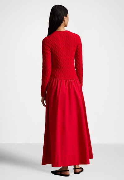 Rdeča dolga obleka z zgornjim delom iz pletenega vzorca in padajočo krilnico, s dolgimi rokavi ter nabranim pasom. Oblečena z ravnimi črnimi sandali.