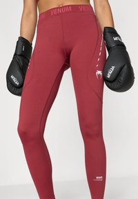 Leggings atléticas cor de vinho com uma cintura larga, apresentando o logótipo Venum branco e detalhes, combinadas com luvas de boxe pretas. Textura suave.