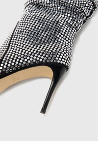 Cizmă neagră cu toc înalt, confecționată dintr-un material texturat acoperit cu nituri argintii, având un vârf ascuțit și un toc stiletto subțire.