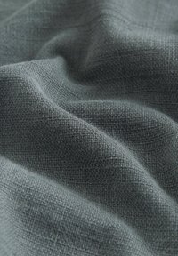 Kuvioitu, kudottu kangas hillityssä harmaassa värissä, jossa on runsas, kosketeltava pinta näkyvine ompeleineen ja pehmeä laskos.