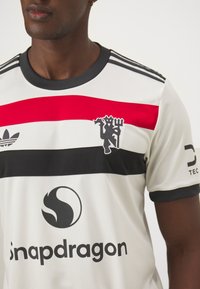 Maglia sportiva bianca con strisce orizzontali nere, rosse e grigie. Presenta un'emblema nero, logo e nome del marchio in lettere grasse.