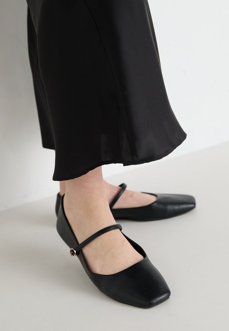 Chaussures plates noires avec un design à bout carré, fabriquées en cuir lisse, dotées d'une seule sangle sur le pied, avec un petit accent décoratif.